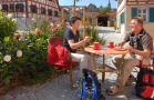 Zwei Wanderer sitzen im Straßencafe bei Kaffee und Kuchen. Im Hintergrund fränkische Fachwerkhäuser.