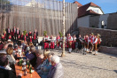 Weinfest auf der Freifläche des neuen Dorfplatzes. Musikgruppe steht, während ein Chor für die Gäste singt. Chor und Musik tragen Tracht.