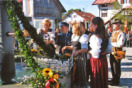 Sechs Personen stehen anlässlich der Einweihung des Dorfplatzes am Brunnen. Im Hintergrund stehen Dorfbewohner und Vereine mit ihren Fahnen.