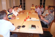 13 Männer sitzen an einem Konferenztisch und beraten sich