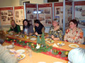 Fröhliche Damen verschiedener Altersklasse beim gemeinsamen Essen. Auf der Tafel: Essensteller, Weihnachtsdeko, Osaft und Sekt. Im Hintergrund Faltaufsteller mit Bauprojektinformationen. 