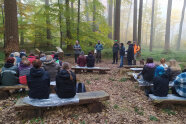 Personen im Wald sitzern auf Holzbänken und lauschen den Vortragenden, die vor ihnen stehen (© Mathias Burghard)