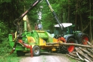 Traktor mit Anhänger zum Zerhacken von Holz. Mittels angebauten Greifarm wird Holz zugeführt.
